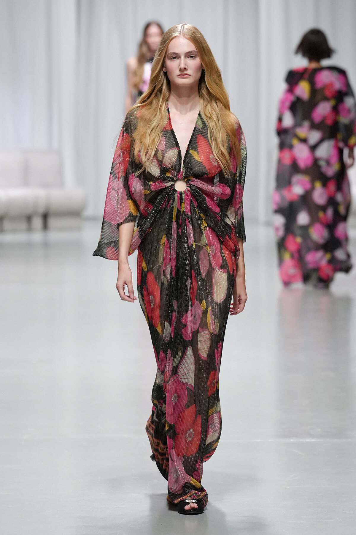 Model walks a runway in a sheer black floral gown with a waist knot and circular cutout at the midriff.