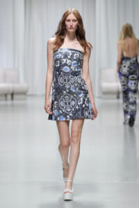 Model walks the runway in a strapless, ornate blue-gray patterned mini dress with white platform sandals.