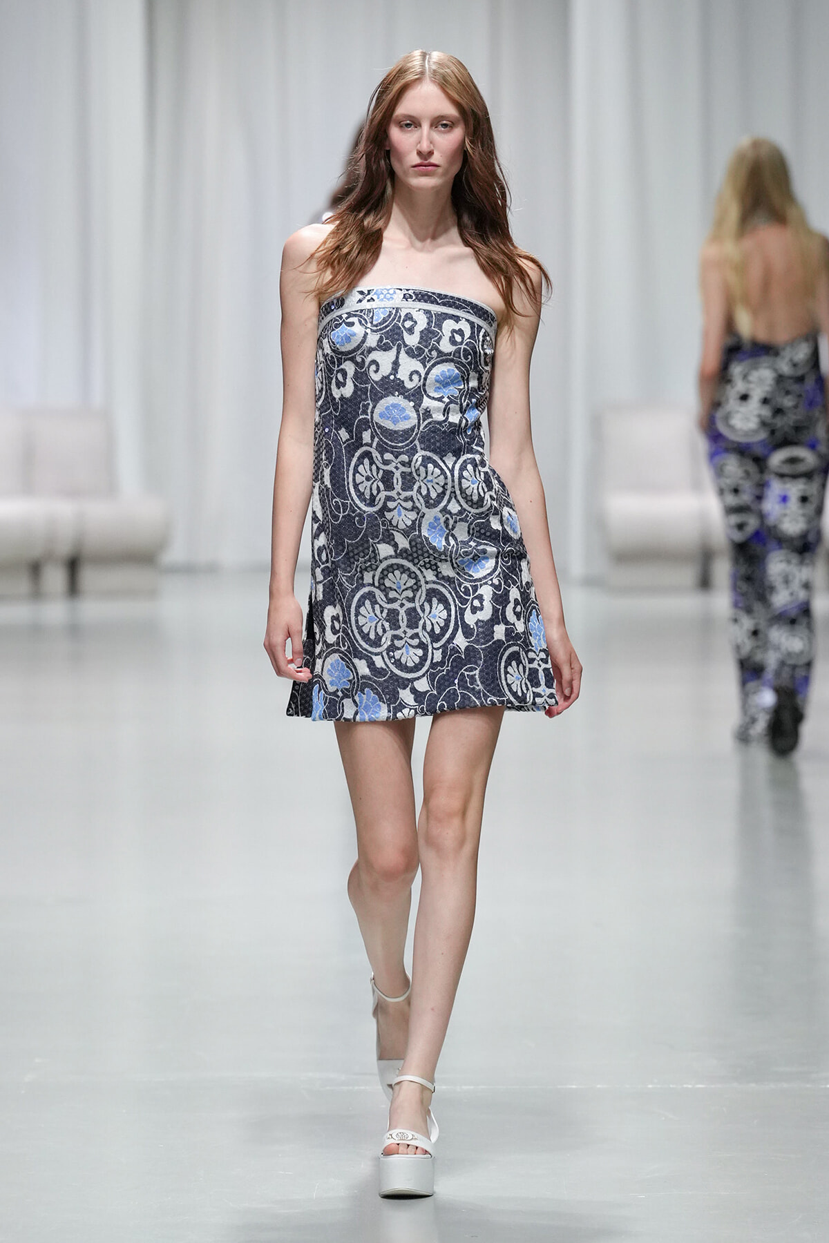 Model walks the runway in a strapless, ornate blue-gray patterned mini dress with white platform sandals.