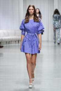 Model walking a fashion runway in a blue geometric-print dress with puff sleeves and a waist tie belt, white platform heels.