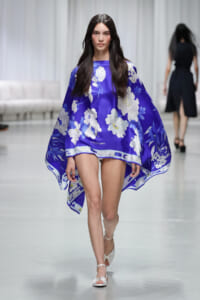 Model walking a fashion show in a vivid purple cape dress with white floral print and white sandals.