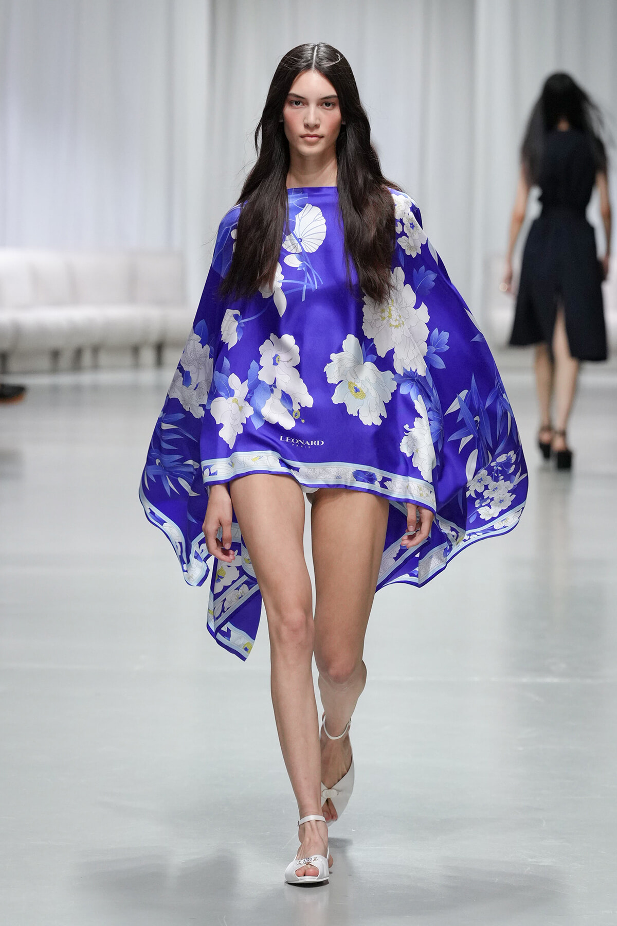 Model walking a fashion show in a vivid purple cape dress with white floral print and white sandals.