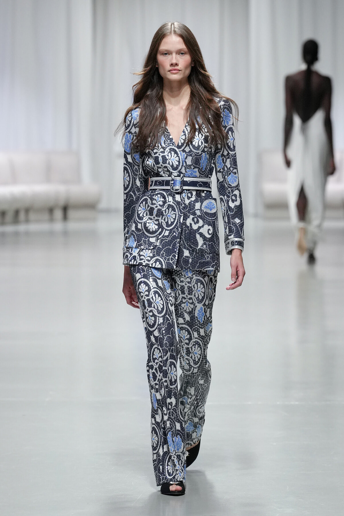 Model walking a fashion show wearing a blue-gray paisley-patterned suit with a matching belt.