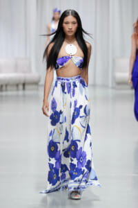 Model walks the runway in a blue and white floral bandeau top with a matching pleated maxi skirt, accessorized by a large circular pendant necklace.