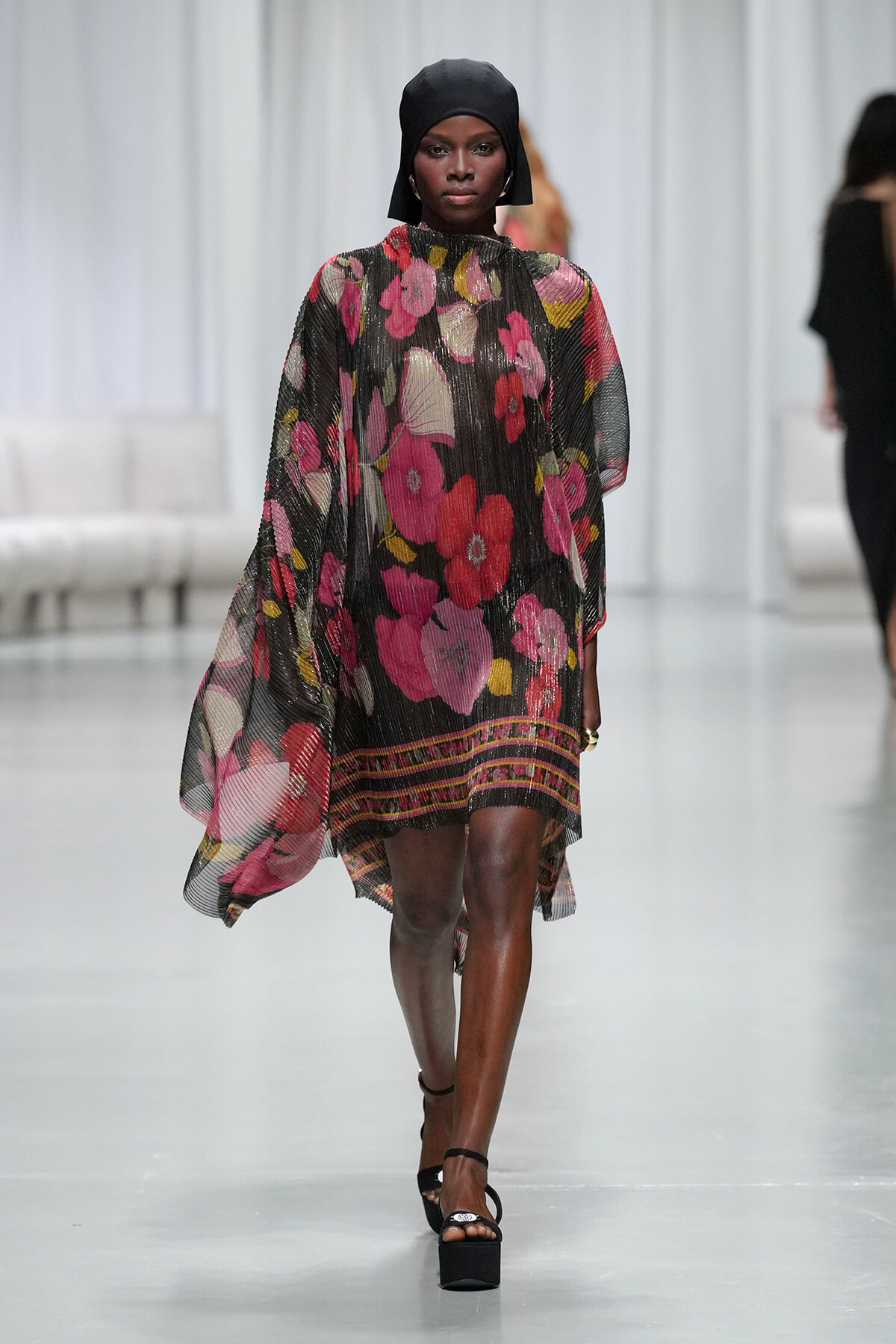 Model walking the runway in a sheer black floral cape, black headwrap, and platform sandals.