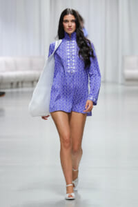 Female model walking a fashion runway in a purple patterned long-sleeve romper with a high collar, white tote over her shoulder, white sandals.