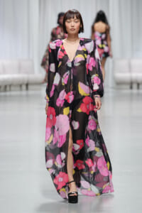Model walks the runway wearing a sheer black floral gown with pink, red, and yellow flowers.