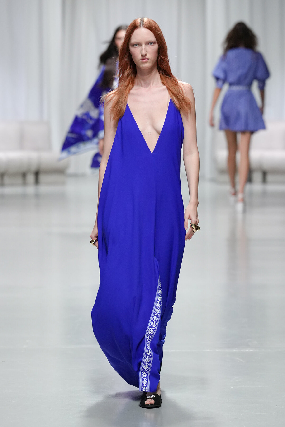 Model walking a runway in a cobalt blue plunging-neckline maxi dress with a white floral trim along the side seam and black sandals.