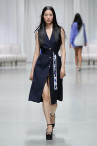 Model walking runway in a navy sleeveless belted trench dress with a high slit and floral-accent belt, pairing with black platform sandals.