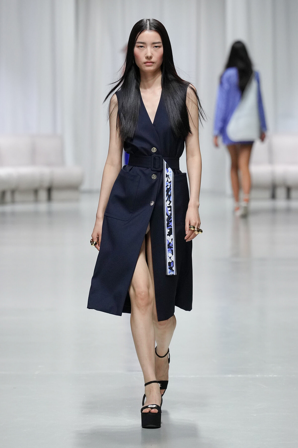 Model walking runway in a navy sleeveless belted trench dress with a high slit and floral-accent belt, pairing with black platform sandals.