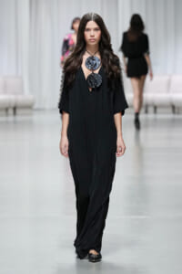 Model walks a minimalist black gown with wide sleeves and two large circular pendant necklaces on a black cord. The jewelry stands out against the outfit as she models on a runway.