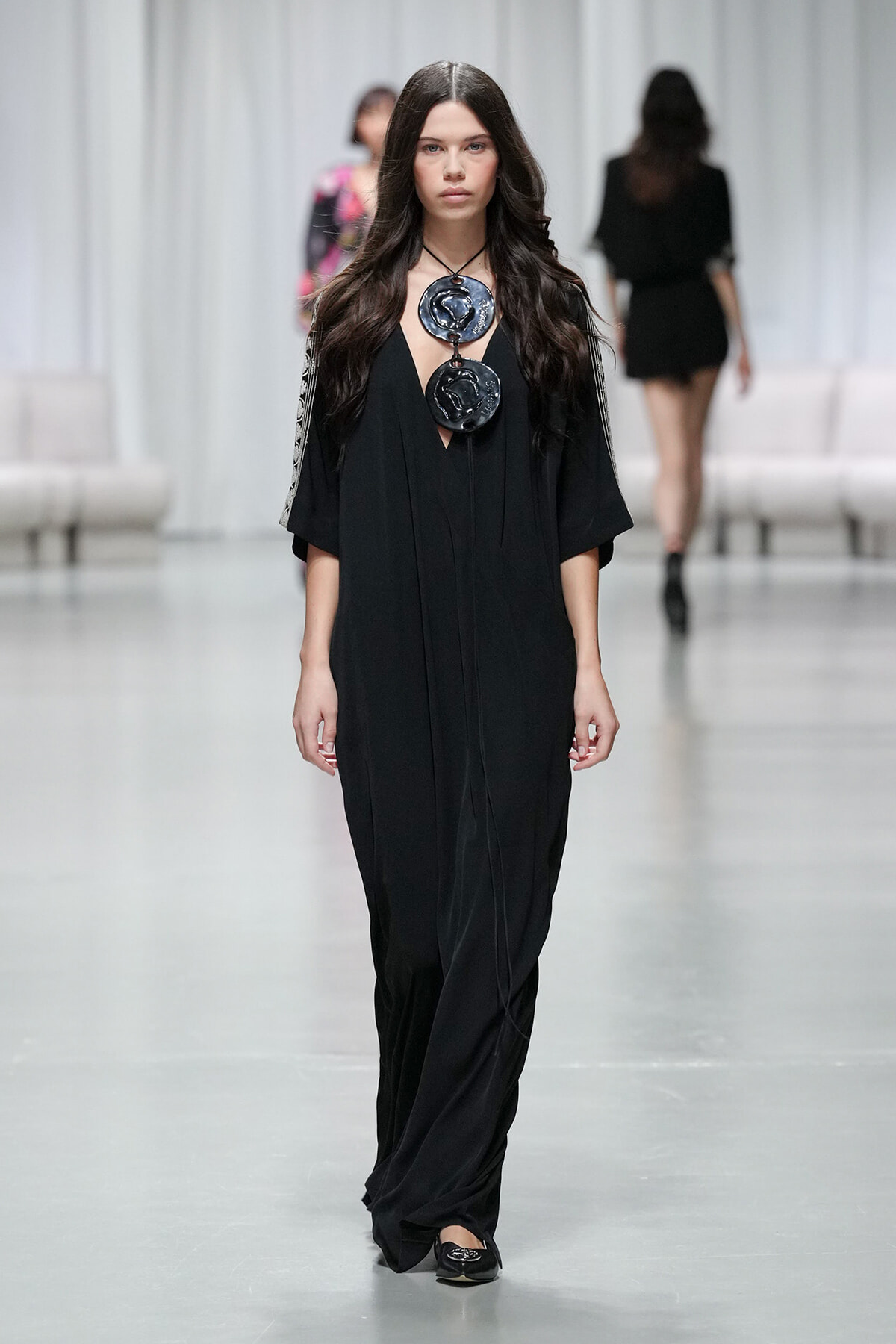 Model walks a minimalist black gown with wide sleeves and two large circular pendant necklaces on a black cord. The jewelry stands out against the outfit as she models on a runway.