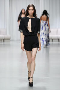 Model walking the runway in a black romper with a deep center cutout, short sleeves with embroidered cuffs, and platform sandals.