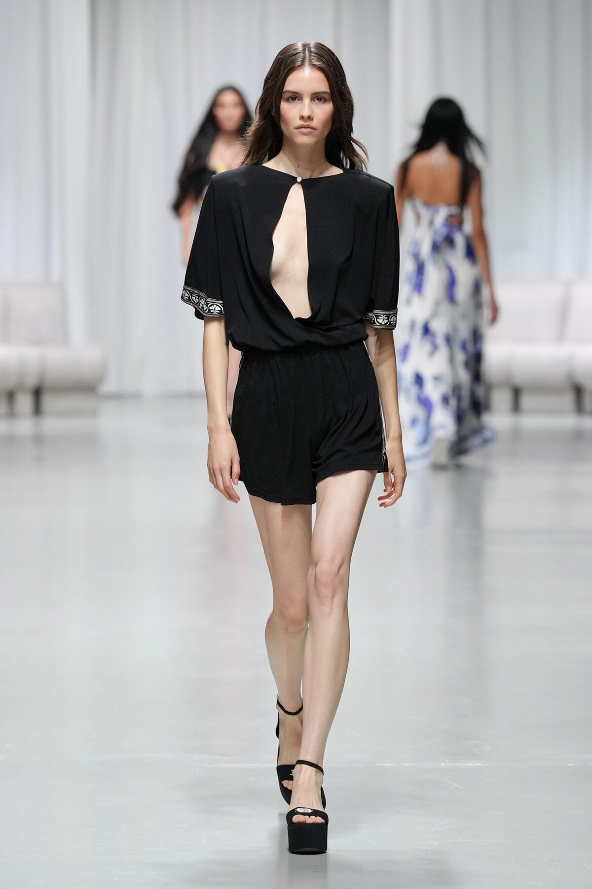 Model walking the runway in a black romper with a deep center cutout, short sleeves with embroidered cuffs, and platform sandals.