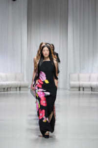 Model walking the runway in a black sleeveless maxi dress with bold pink and purple floral print.