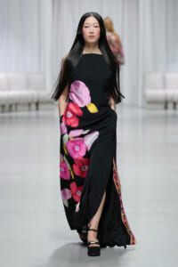 Model walking a fashion show in a black maxi dress with large pink and magenta flowers on the skirt.