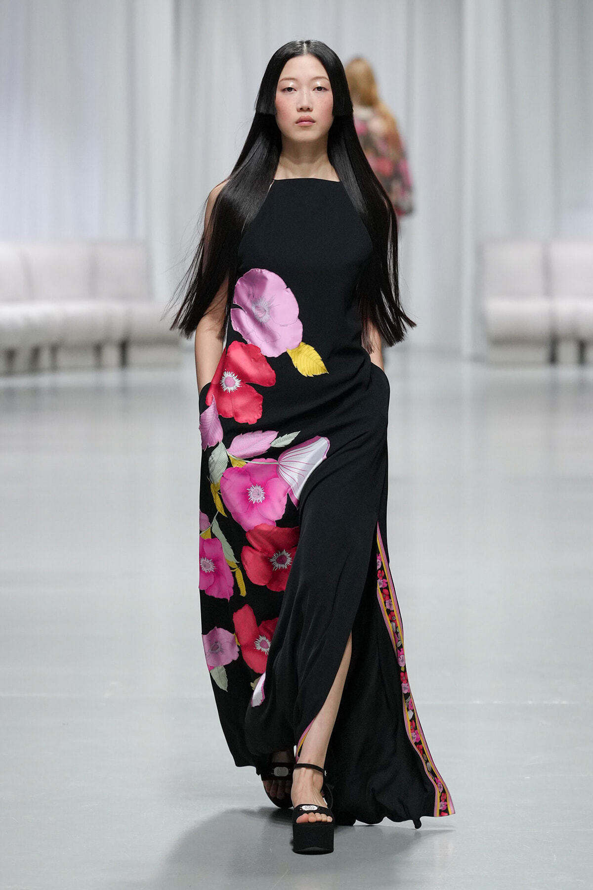 Model walking a fashion show in a black maxi dress with large pink and magenta flowers on the skirt.