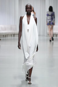 Model walking a fashion show runway in a flowing white halter gown and white sandals, minimalist styling.