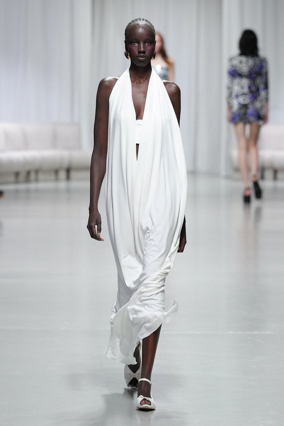 Model walking a fashion show runway in a flowing white halter gown and white sandals, minimalist styling.
