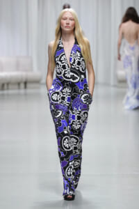 Model walking the runway in a black, white and purple floral halter jumpsuit with pockets atop a light-gray catwalk.