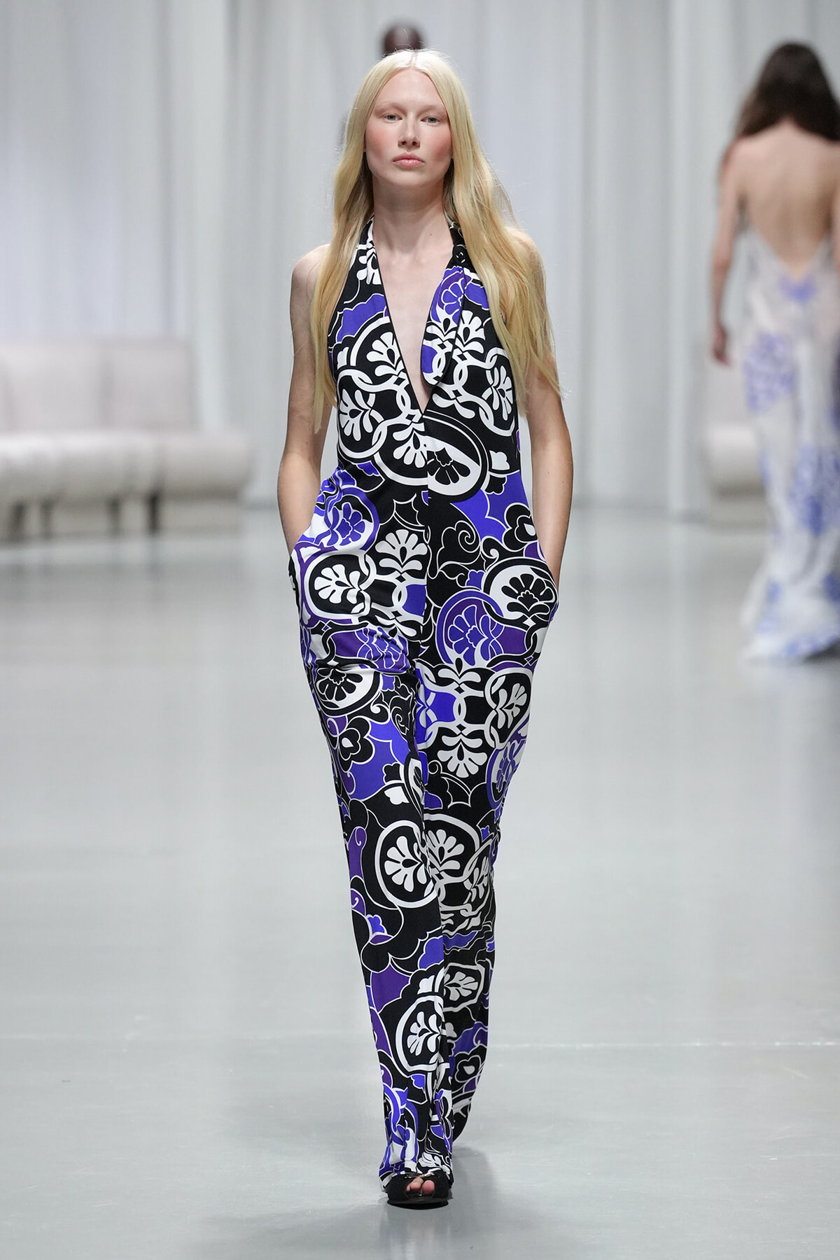 Model walking the runway in a black, white and purple floral halter jumpsuit with pockets atop a light-gray catwalk.