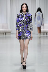 Model walks the runway in a purple, black, and white patterned mini dress with three-quarter sleeves and black platform sandals.
