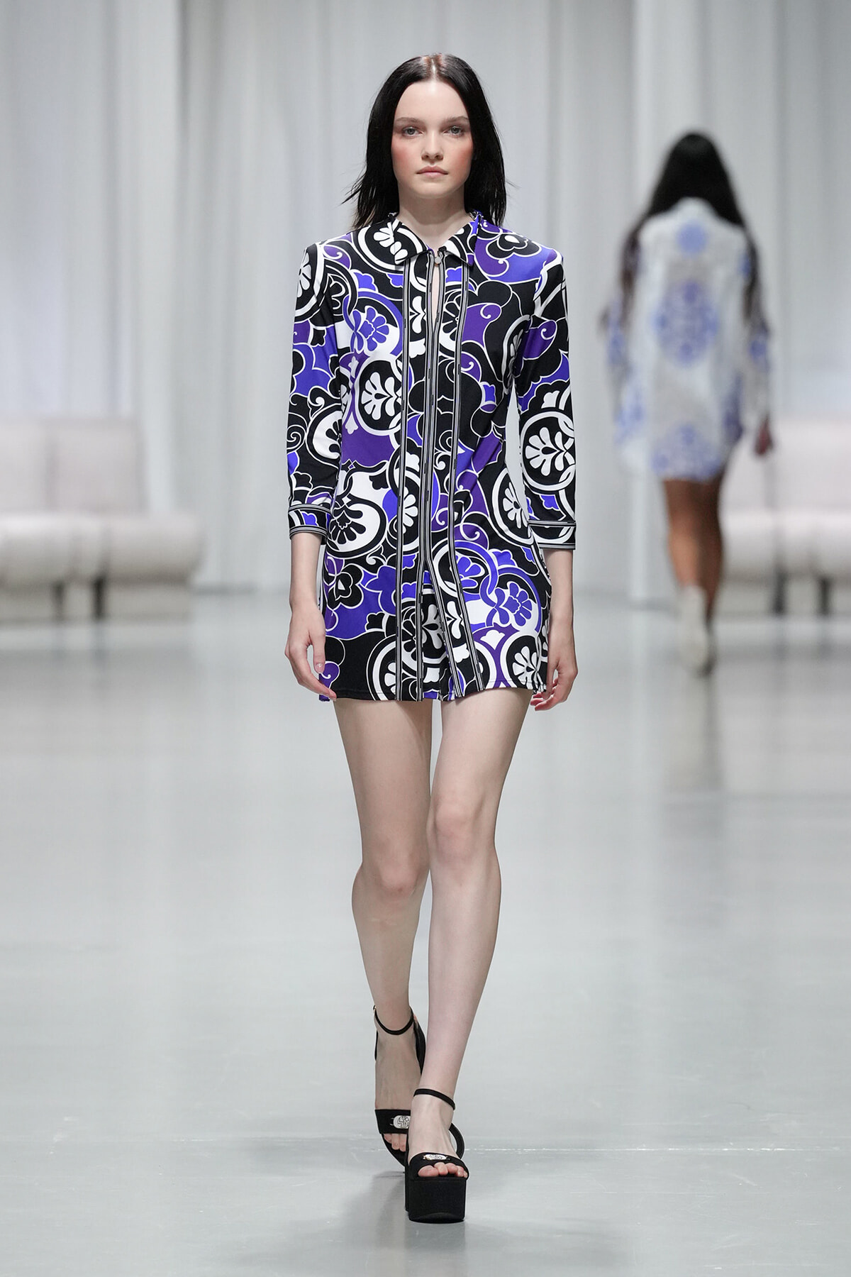 Model walks the runway in a purple, black, and white patterned mini dress with three-quarter sleeves and black platform sandals.