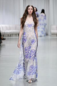 Model walks the runway in a floor-length white dress with purple geometric patterns.