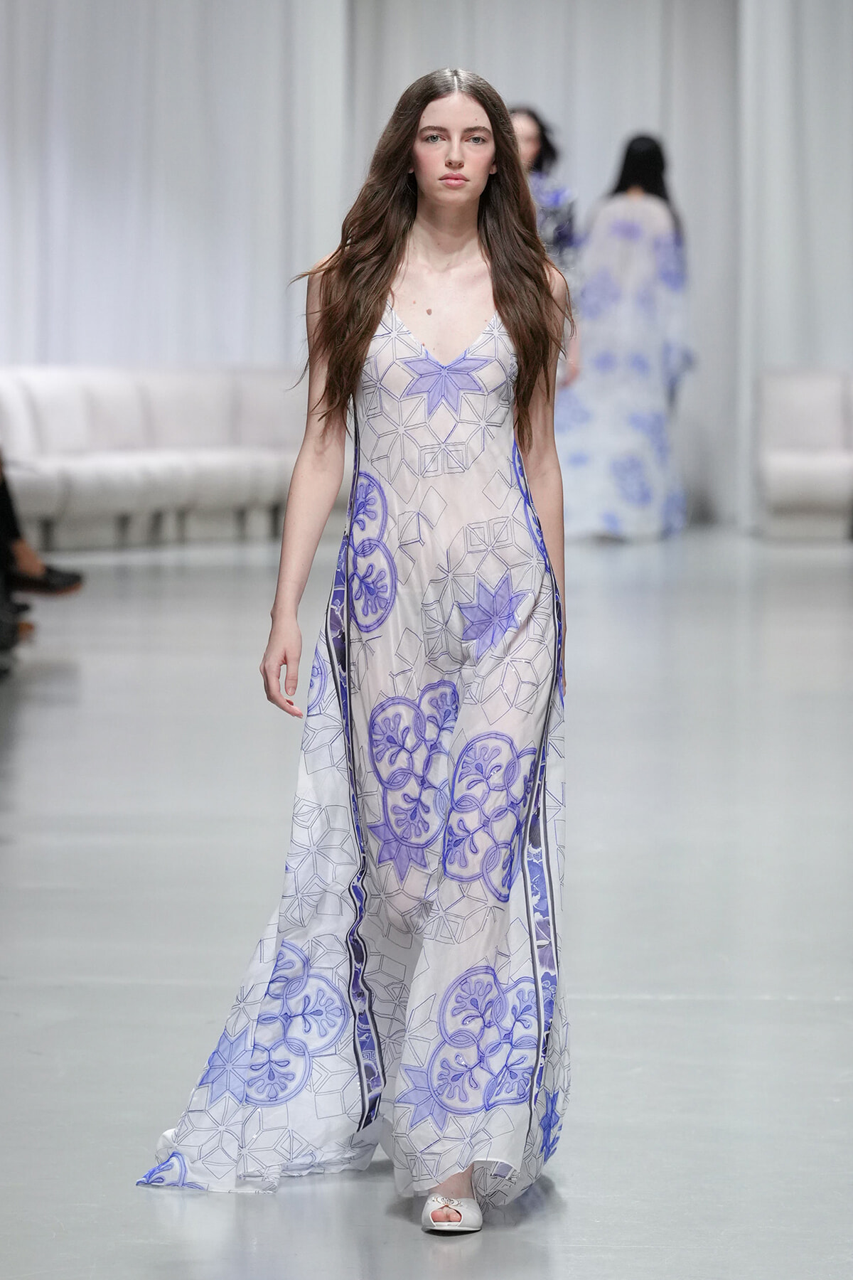 Model walks the runway in a floor-length white dress with purple geometric patterns.
