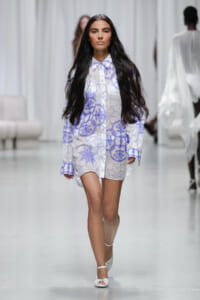 Model walks a runway in a white shirt dress with lavender geometric and floral pattern, long dark hair flowing behind her.