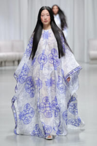 Runway model wearing a flowing white cape with lavender geometric and circular floral prints on the catwalk.