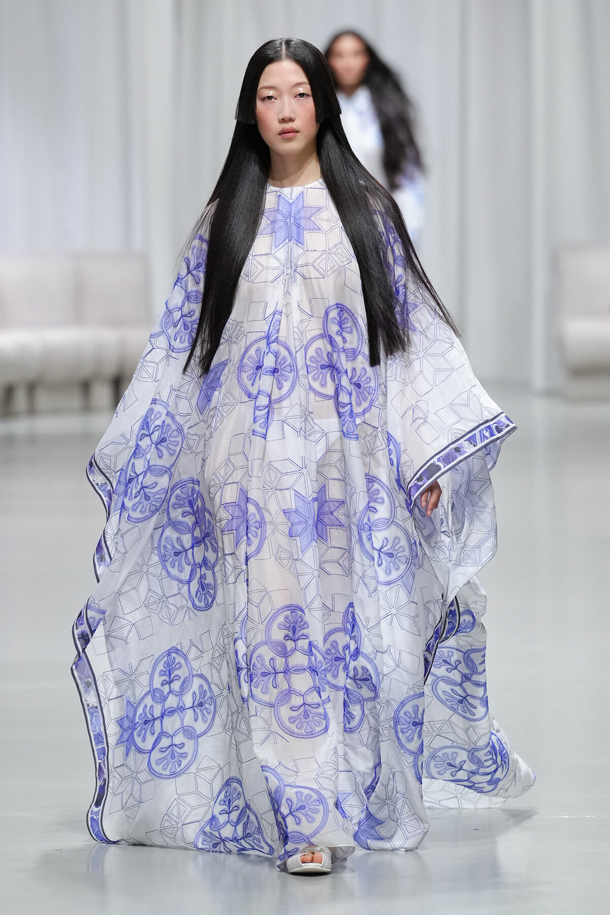 Runway model wearing a flowing white cape with lavender geometric and circular floral prints on the catwalk.