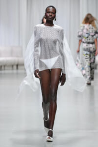 Model walks the runway in a sheer white cape layered over a white bikini, accessorized with large hoop earrings.