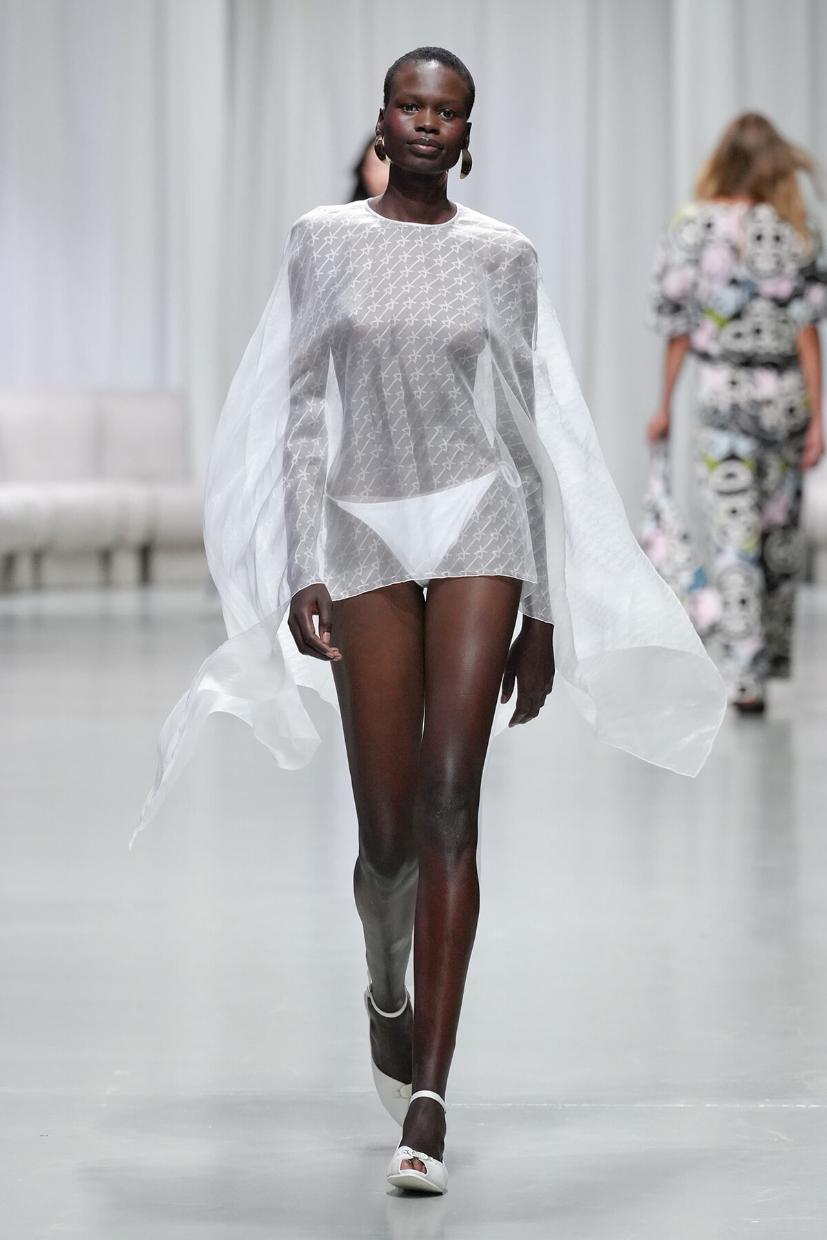 Model walks the runway in a sheer white cape layered over a white bikini, accessorized with large hoop earrings.