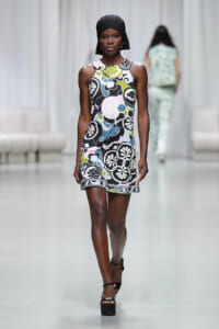 Model walking a runway in a sleeveless multicolored floral mini dress with a black headscarf and platform heels in a fashion show.