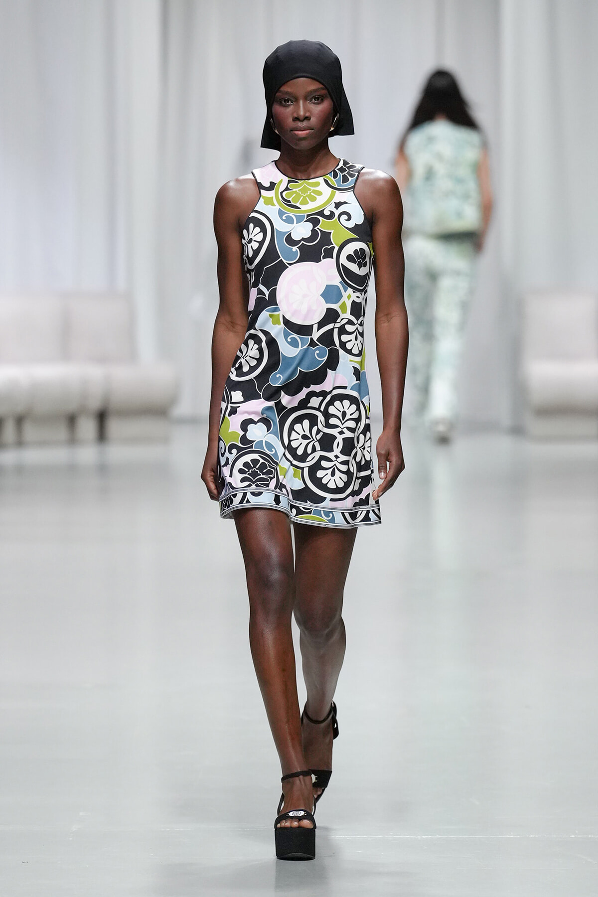 Model walking a runway in a sleeveless multicolored floral mini dress with a black headscarf and platform heels in a fashion show.