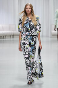 Model walking a fashion show wearing a bold floral-print wide-leg jumpsuit in black, white, pink, blue, and green with a circular neckline detail.