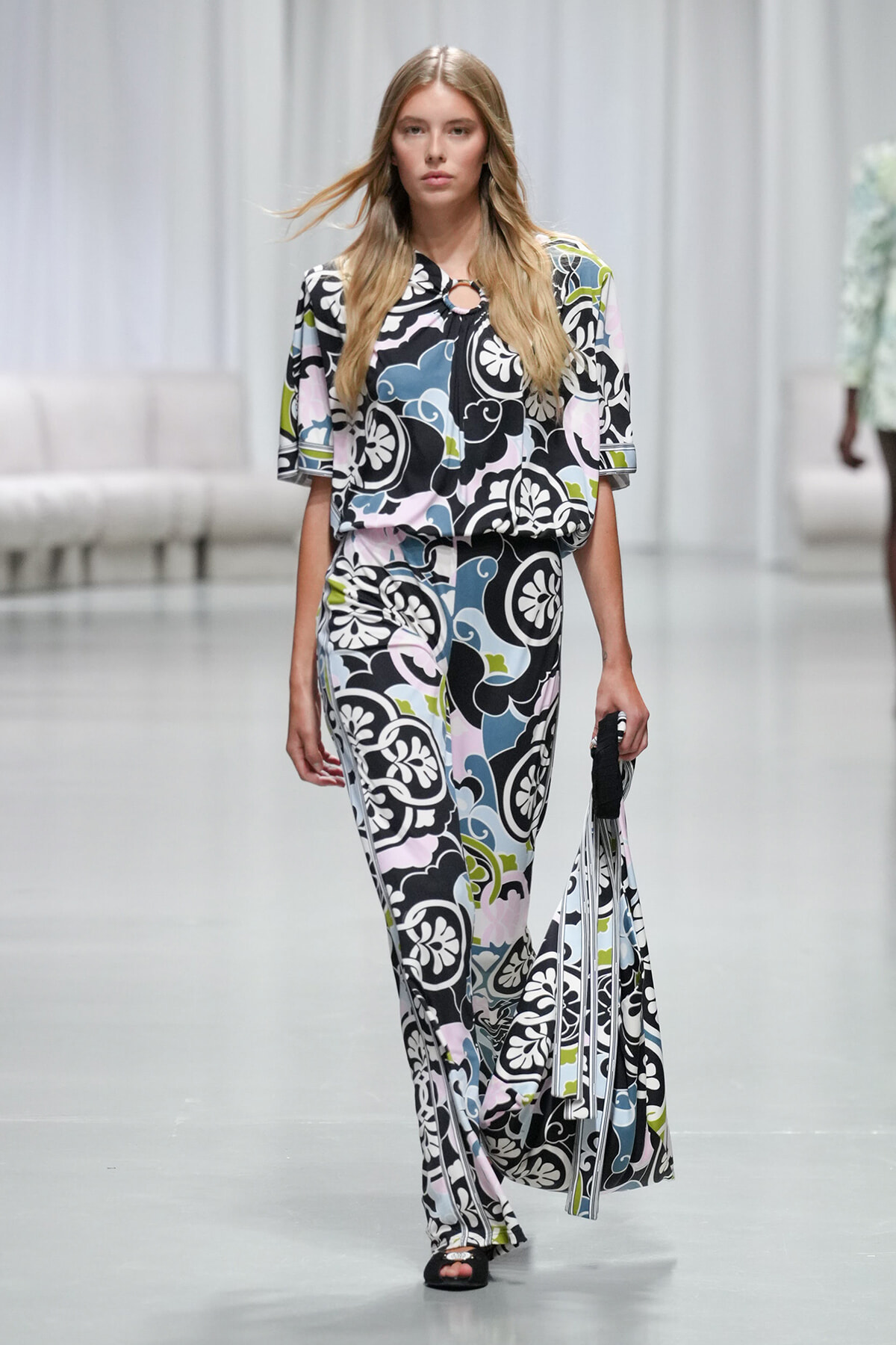 Model walking a fashion show wearing a bold floral-print wide-leg jumpsuit in black, white, pink, blue, and green with a circular neckline detail.