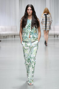 Model walks down a runway in a white and green floral pantsuit with a matching sleeveless vest, hands in pockets.