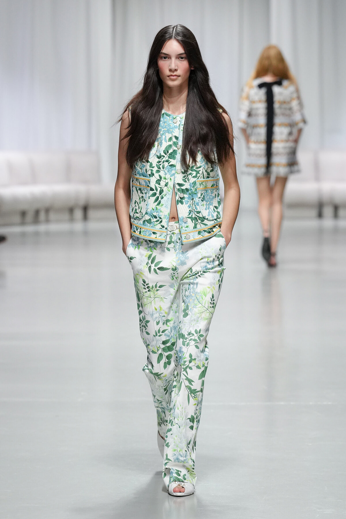 Model walks down a runway in a white and green floral pantsuit with a matching sleeveless vest, hands in pockets.