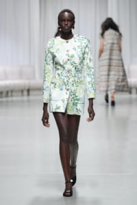 Model walks the runway in a white-green botanical print belted coat with gold buttons and patch pockets.