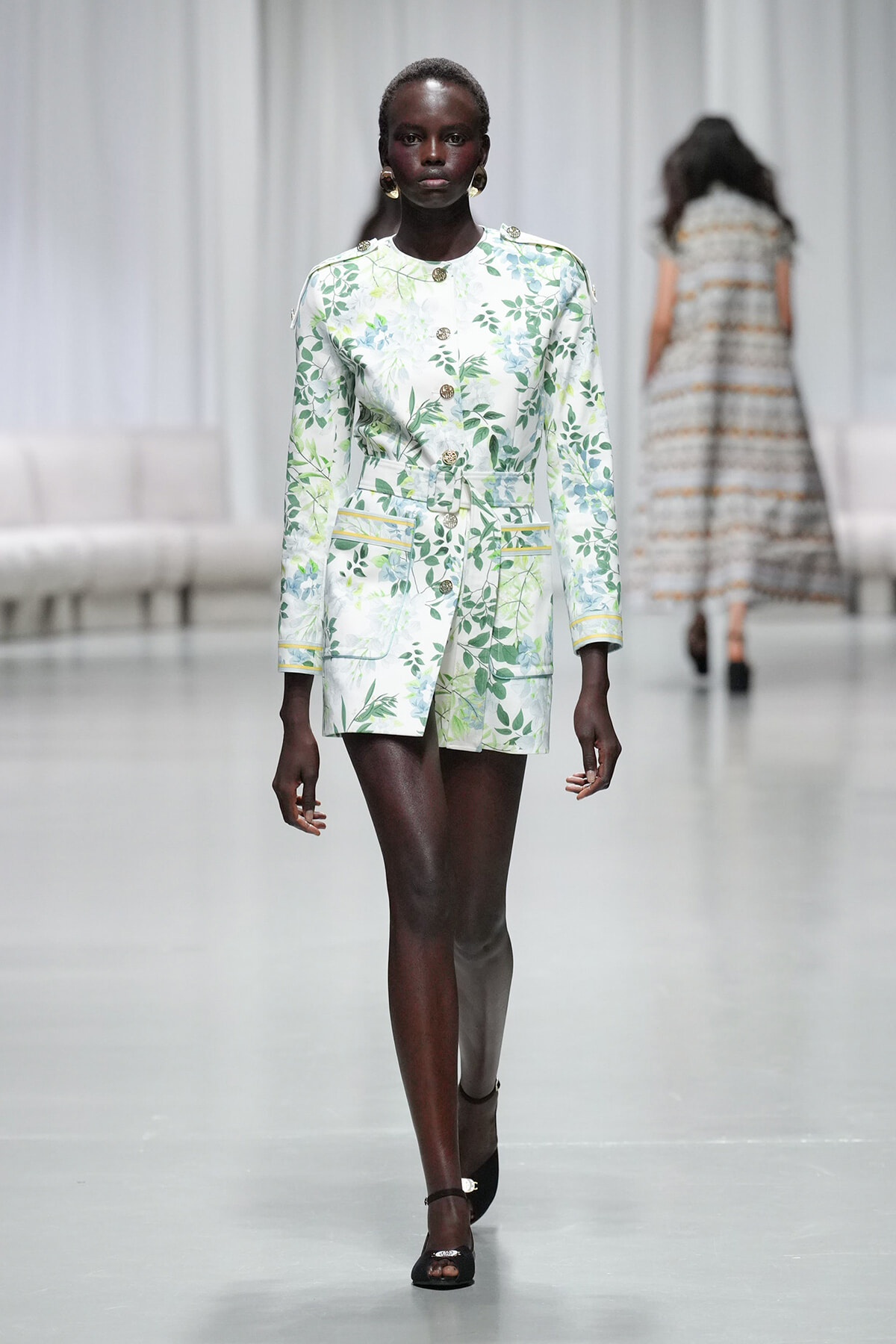 Model walks the runway in a white-green botanical print belted coat with gold buttons and patch pockets.