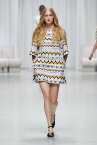 Model walks down runway in a short geometric patterned dress with yellow, gray, and brown zigzag motifs.