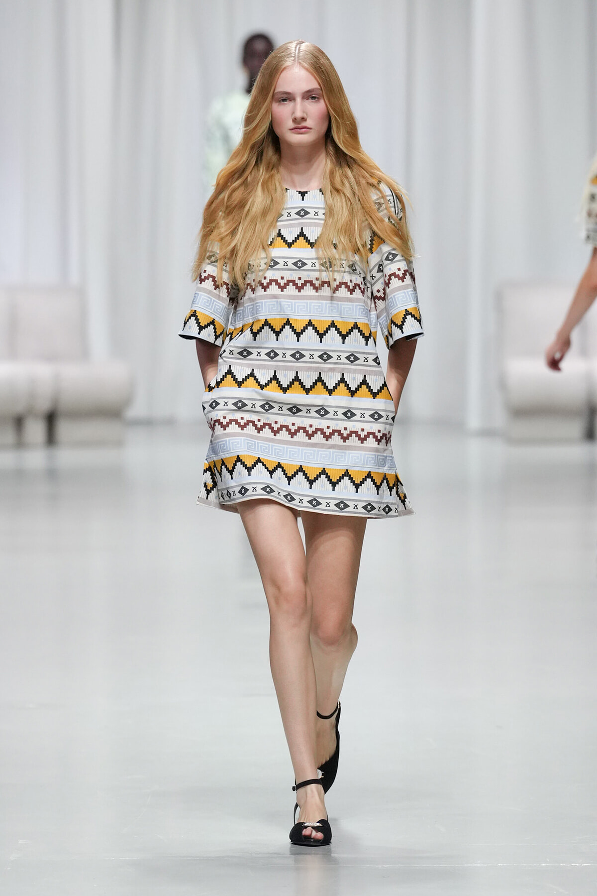 Model walks down runway in a short geometric patterned dress with yellow, gray, and brown zigzag motifs.