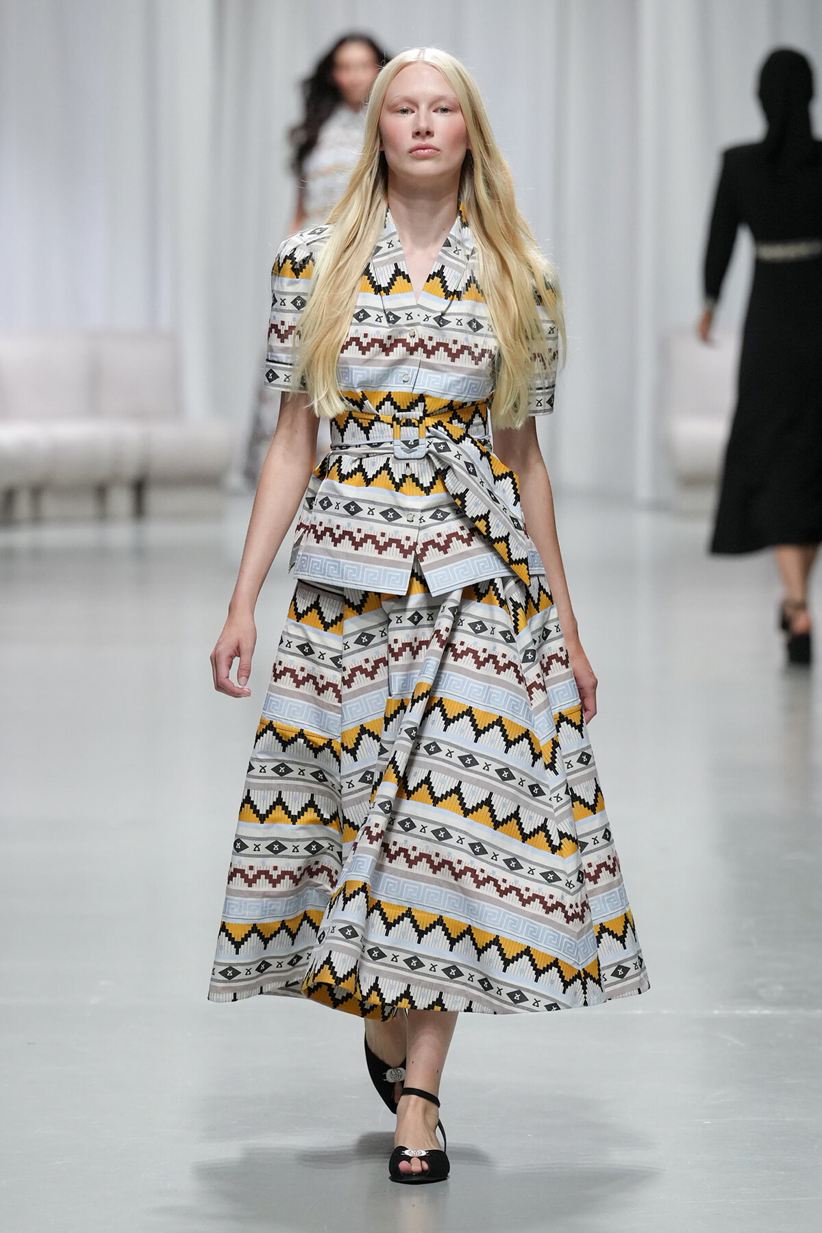Runway model walks wearing a geometric patterned belted midi dress in white, yellow, gray and black with black sandals.