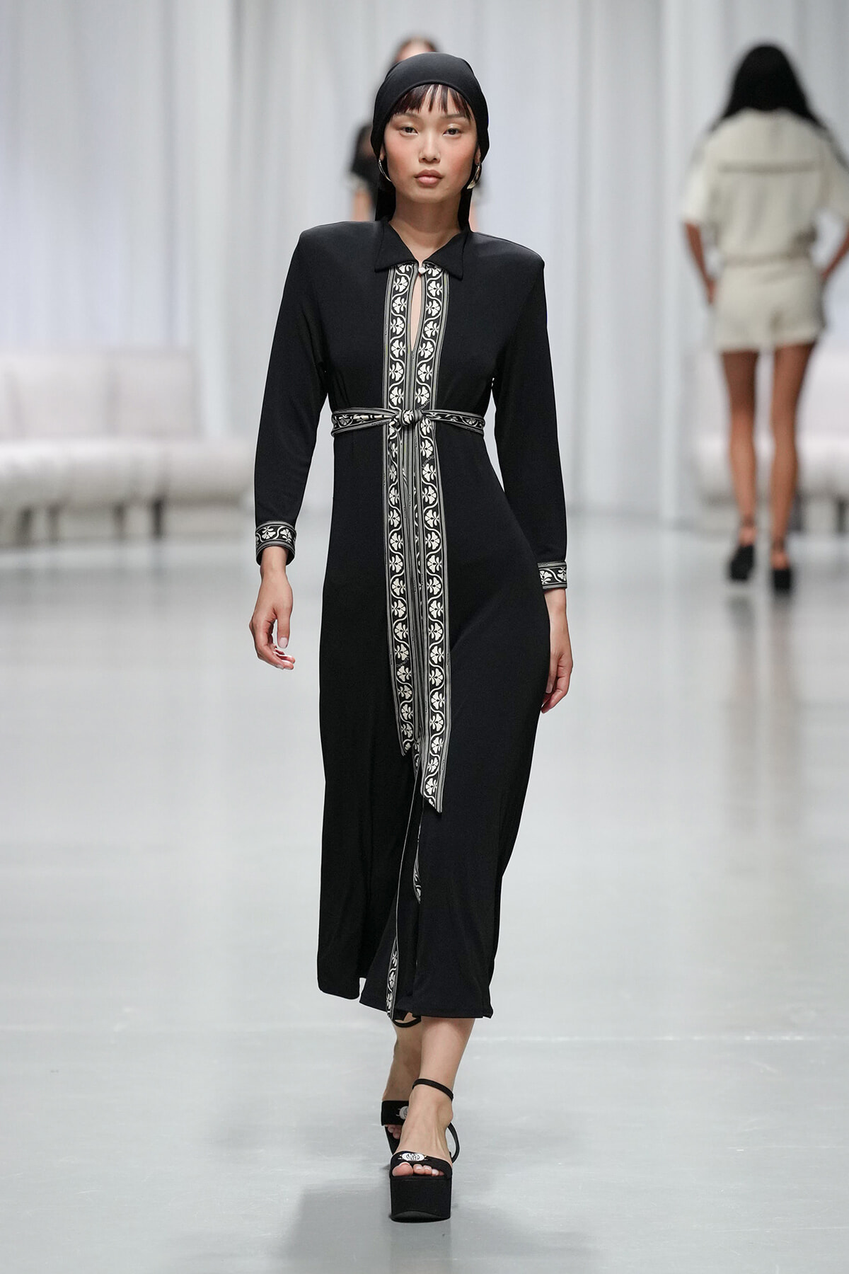 Model walking a runway in a black, long-sleeve dress with white decorative trim and tie belt, paired with a black headscarf and platform sandals