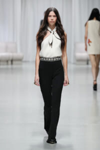 Model walking the runway in a cream halter top with a bow at the neckline and black high-waisted pants with a decorative belt.