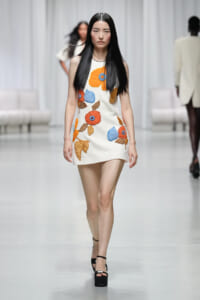 Model walking runway in a white sleeveless mini dress with large orange, blue, and brown floral embroidery.