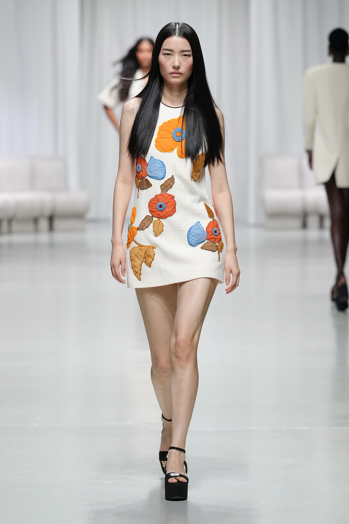 Model walking runway in a white sleeveless mini dress with large orange, blue, and brown floral embroidery.