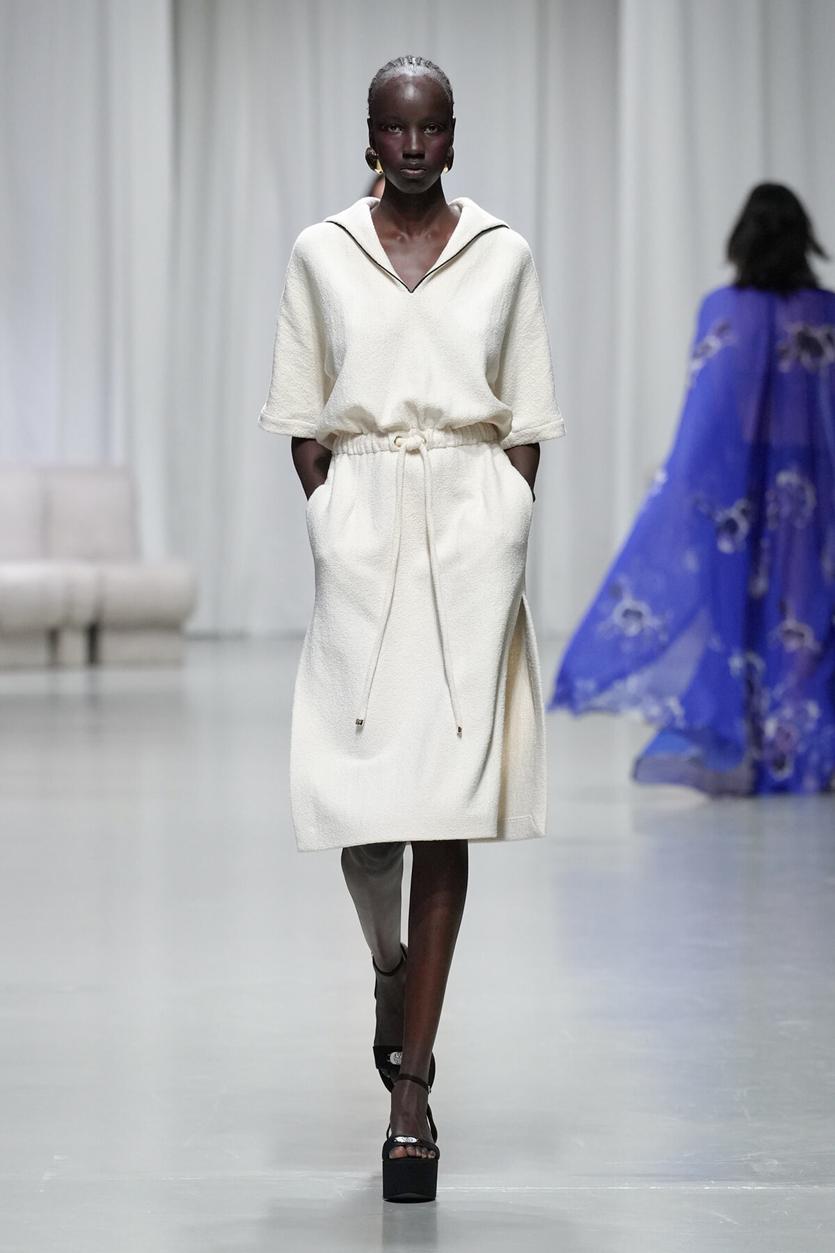 Black female model walks a fashion runway in a white belted knit dress with short sleeves and pockets, paired with black platform sandals.